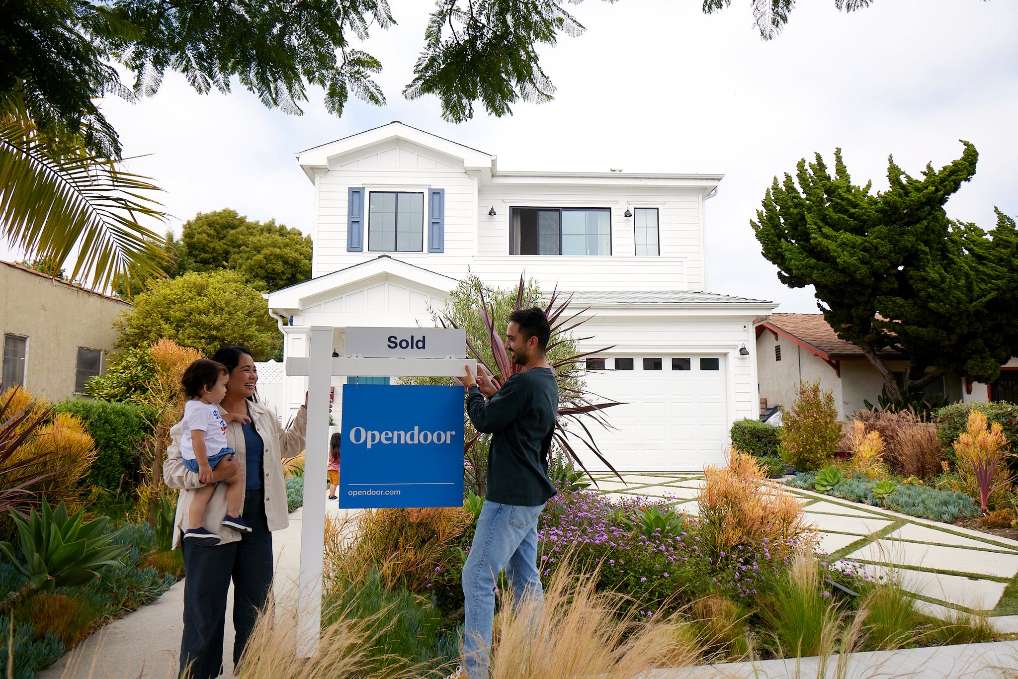 A family stands in front of a white two-story home while a man hangs an Opendoor Sold sign on a signpost.
